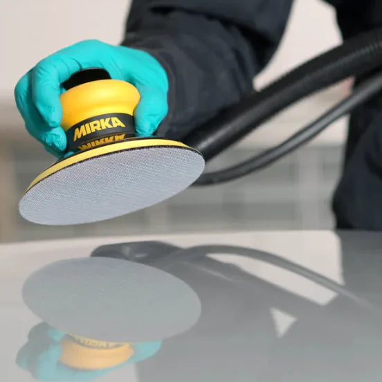 A person wearing a teal glove uses a Mirka 120 grit sanding disc on a glossy white surface, with the Mirka brand name clearly visible on the yellow and black sanding tool.