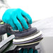 A close-up of a person wearing teal gloves operating a black orbital sander on a light gray surface, with a white protective suit visible in the background.
